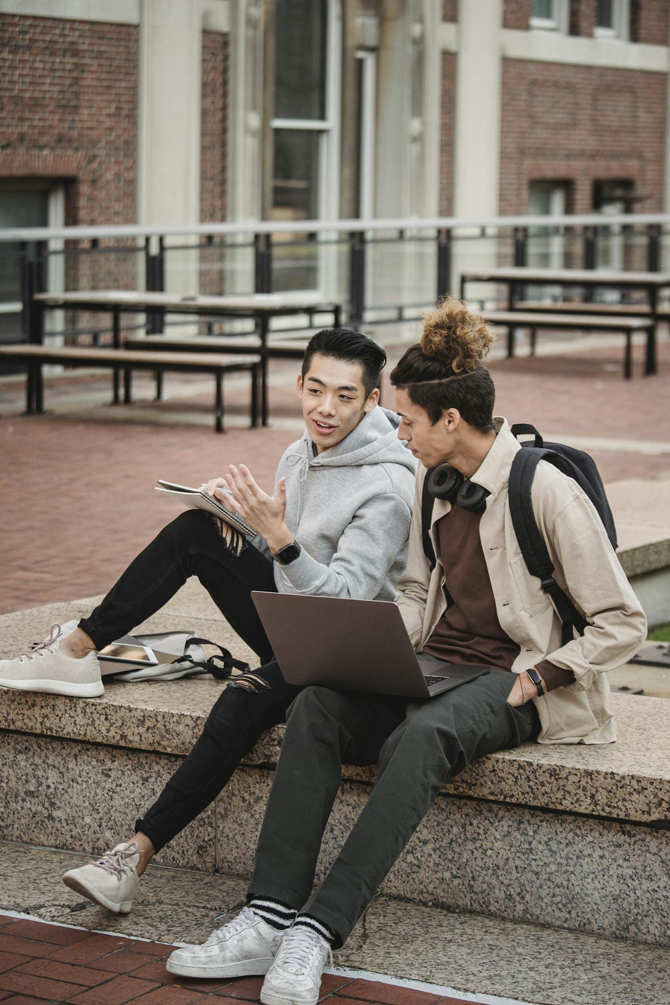 Two young men engage in a lively discussion while using laptops in an outdoor urban setting.